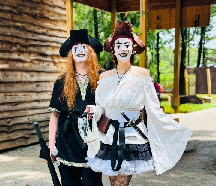 Two costumed people posing outdoors beside a wooden cabin, wearing pirate-style outfits and face paint.