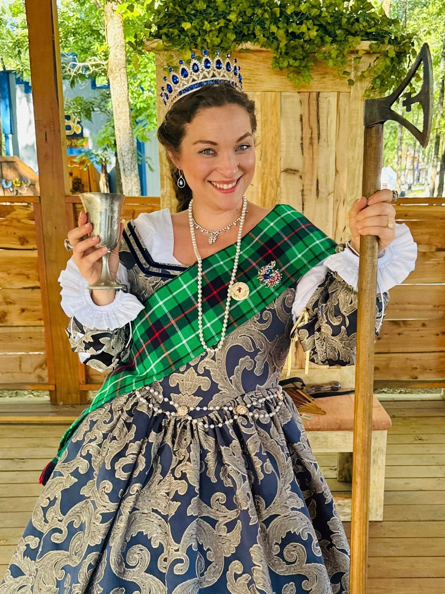 Woman in a blue dress with green sash, smiling and holding a staff in a garden setting