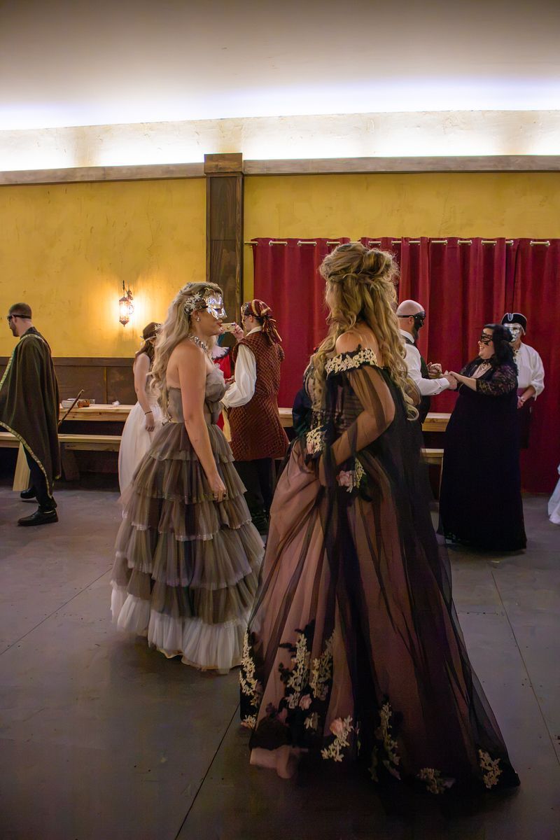 Two women in ornate dresses dance in a hall with red curtains and warm lighting.