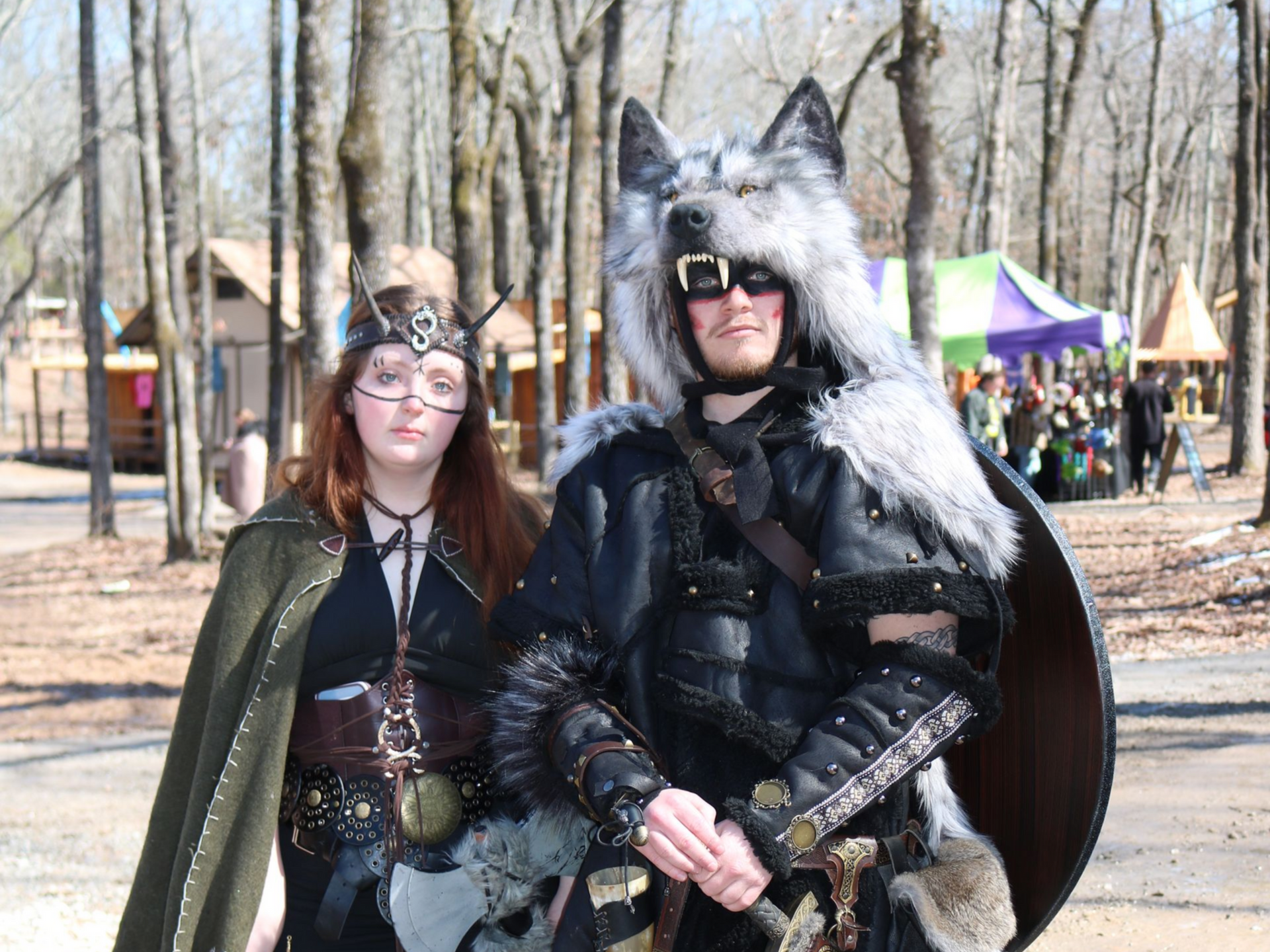 Two people in medieval costumes at an outdoor event. Man wears a wolf head headdress; woman has a cape.