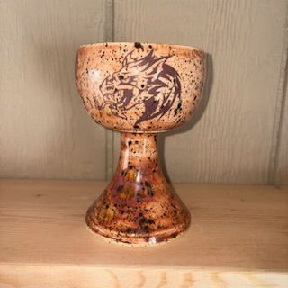 Ceramic goblet with a dragon design. Orange, brown, and black speckled glaze. On a wooden surface.