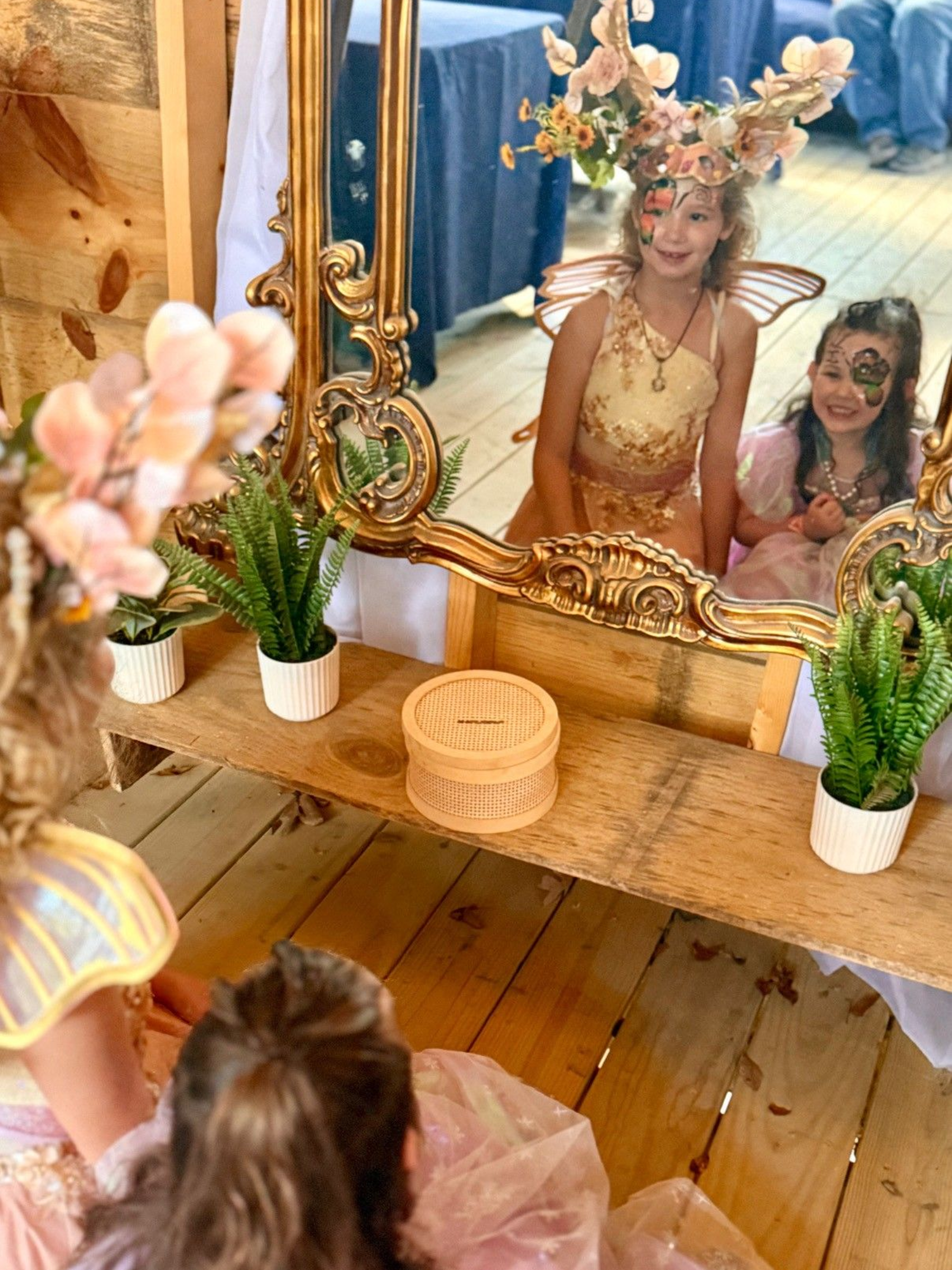 Two girls in fairy costumes pose in front of a mirror with floral decorations.