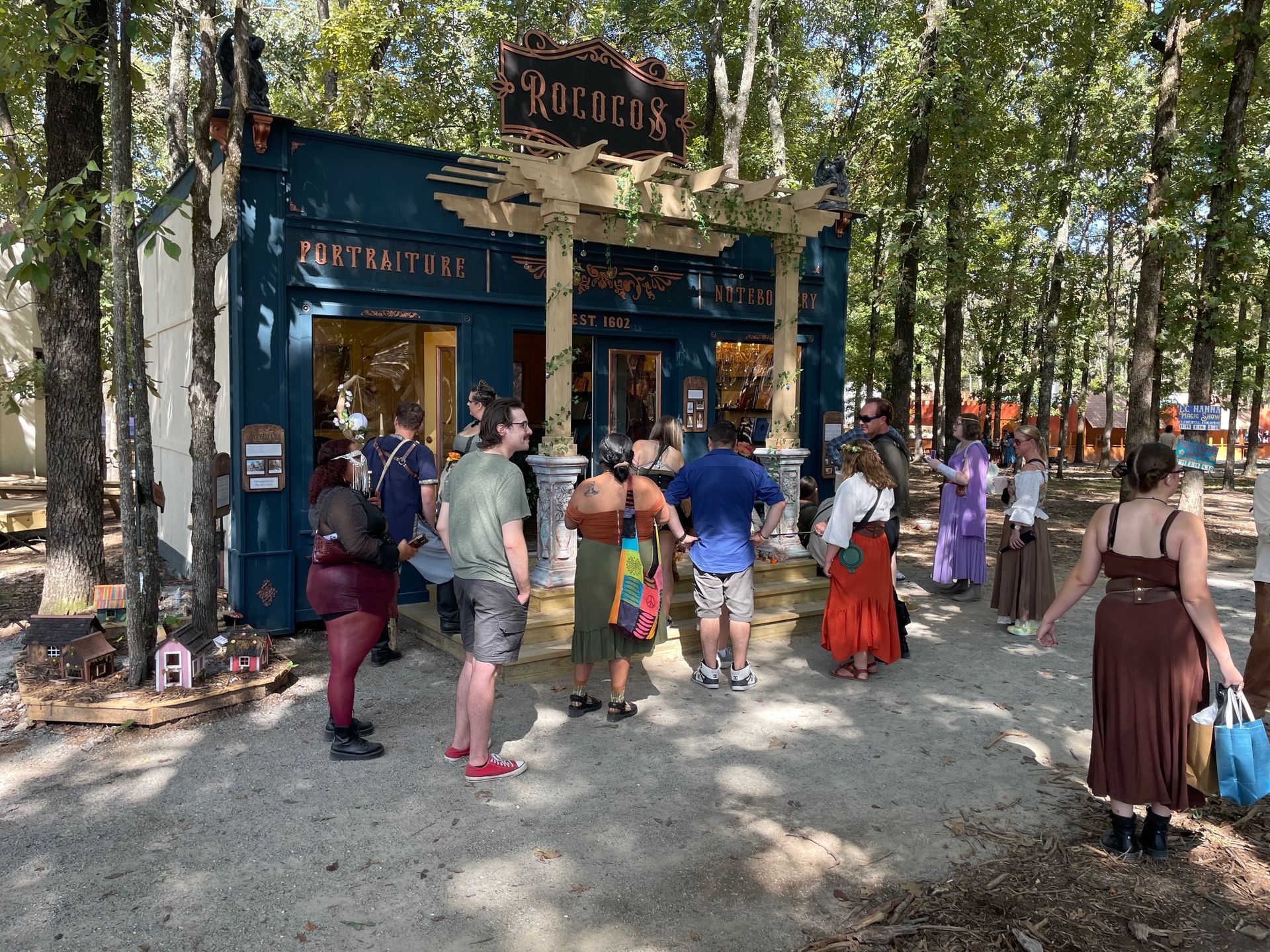 People queue outside a shop with the sign "ROCOCO". Exterior shot, trees in the background. Sunny day.