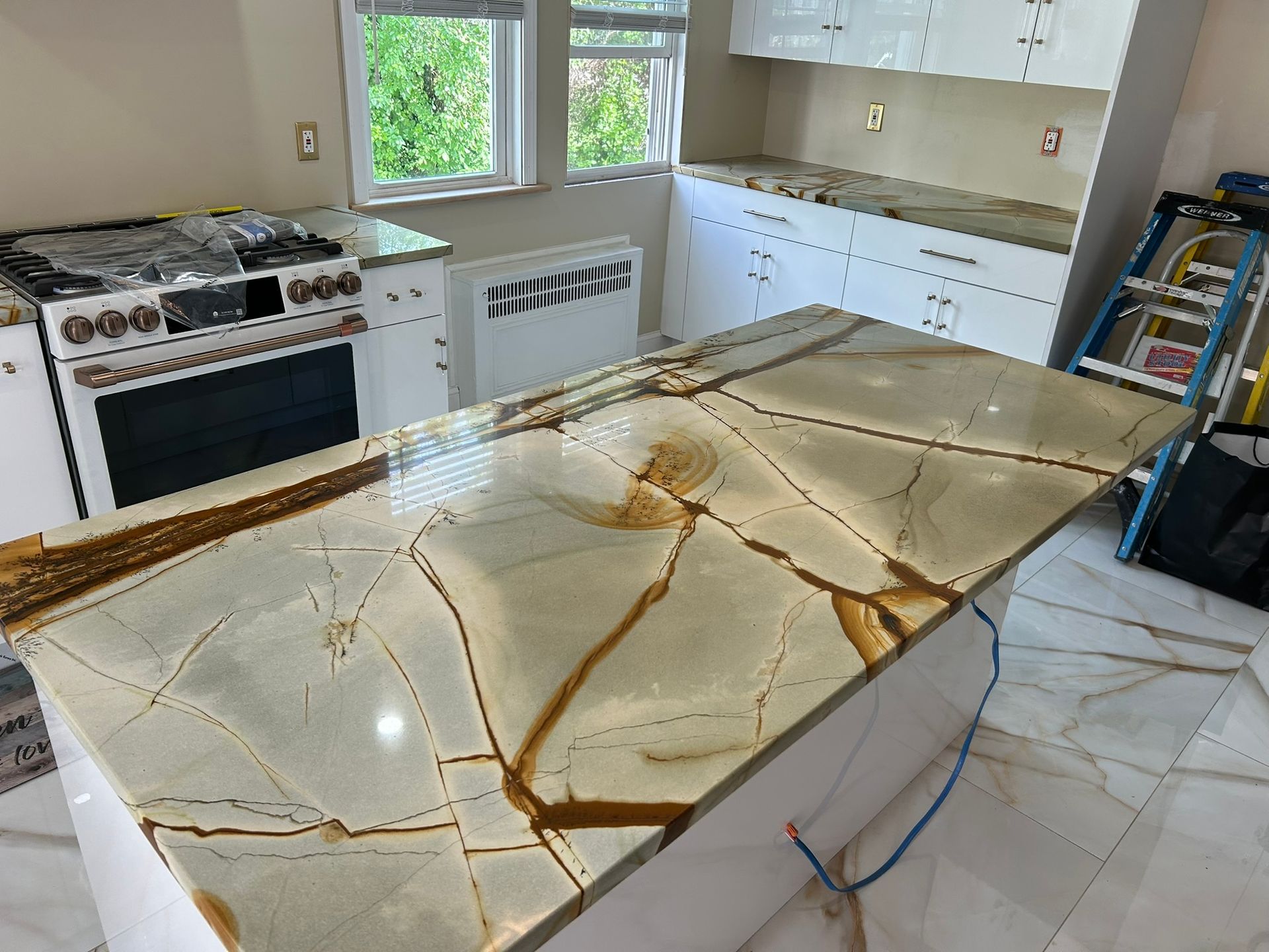 Kitchen with large glossy stone slab on a worktable, cabinets, stove, and window in the background