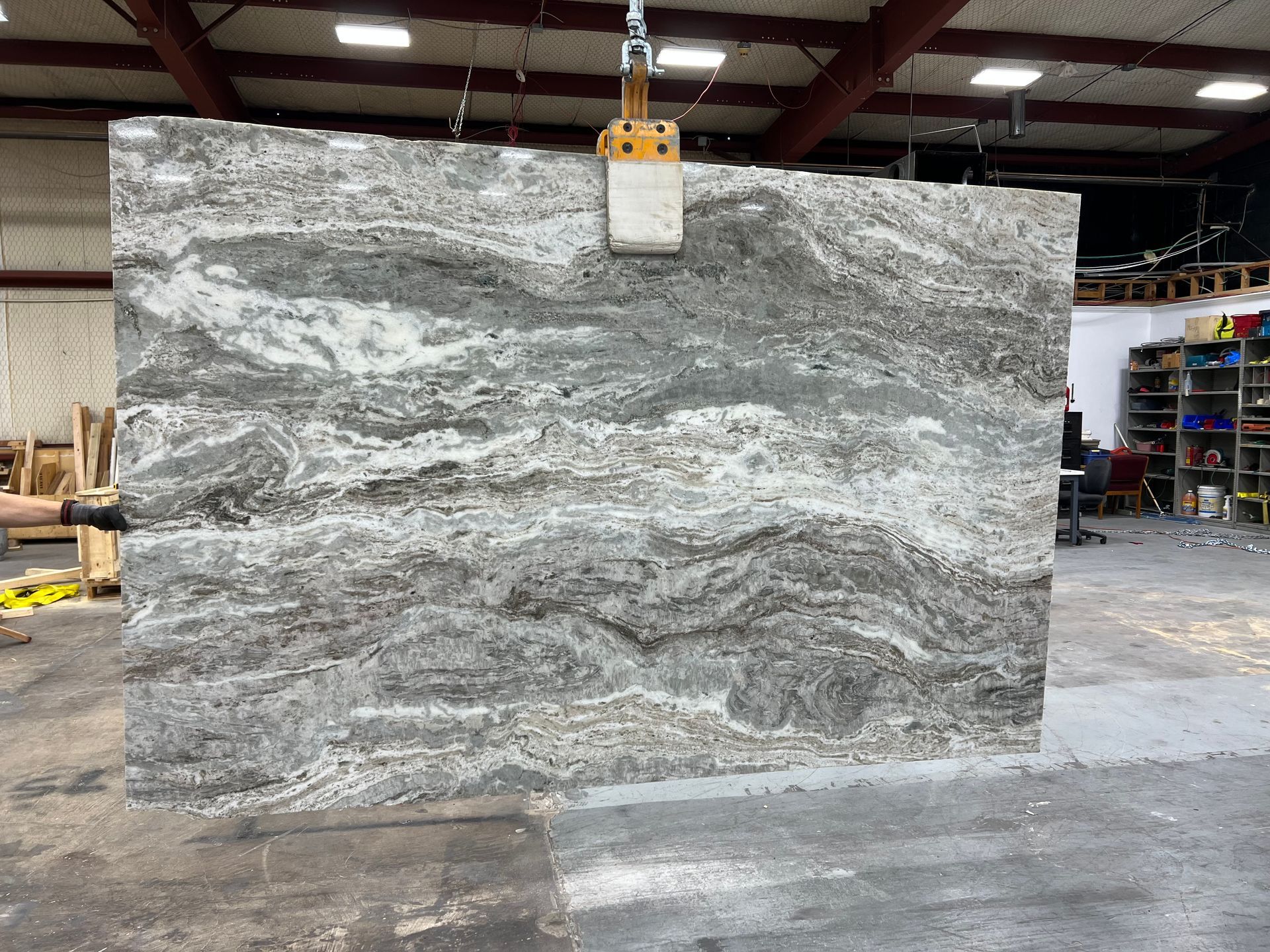 Large granite slab being lifted by a crane in a warehouse, showing gray and white swirling patterns.