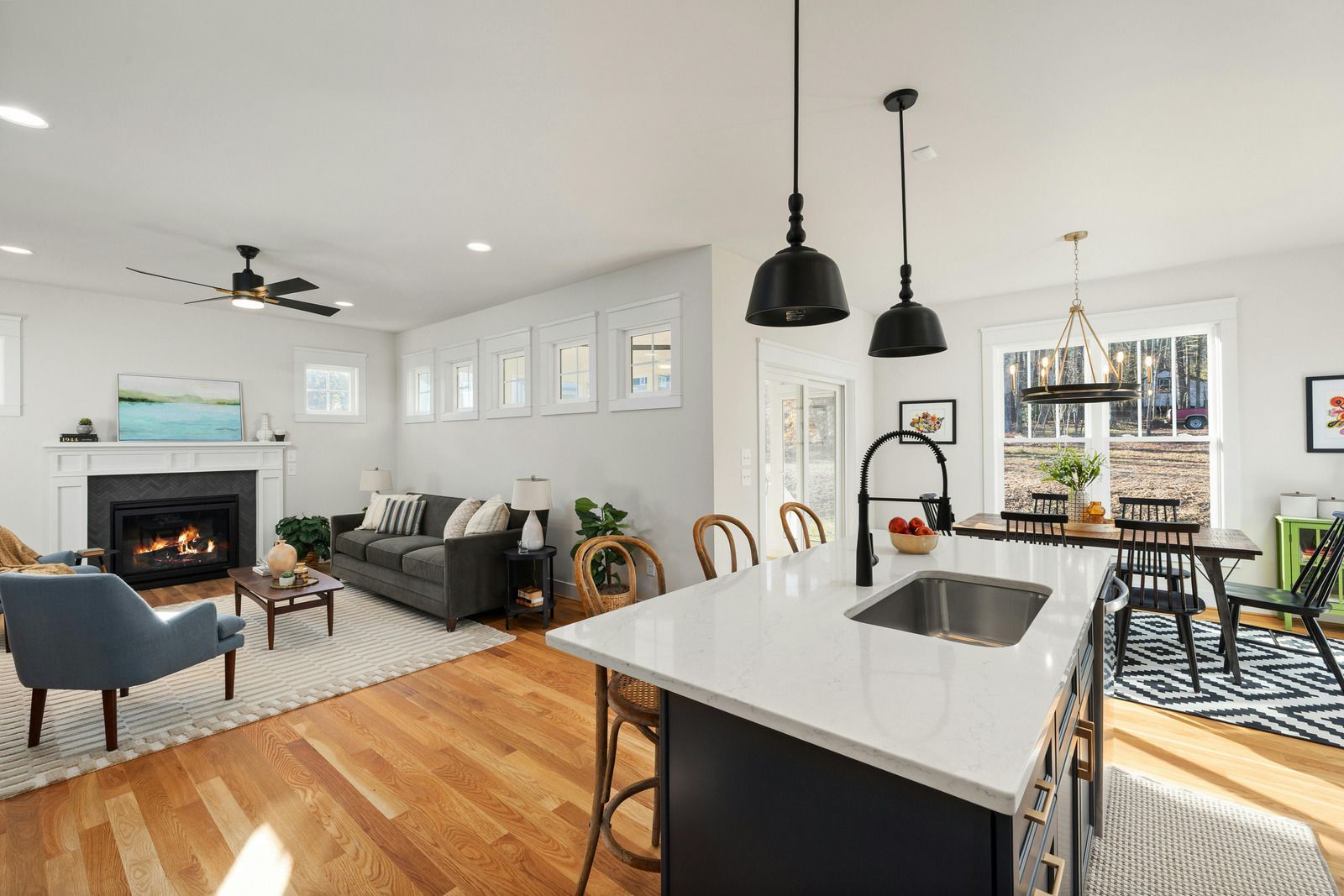 Bright open-plan living room and kitchen with gray sofas, fireplace, pendant lights, and hardwood floors.