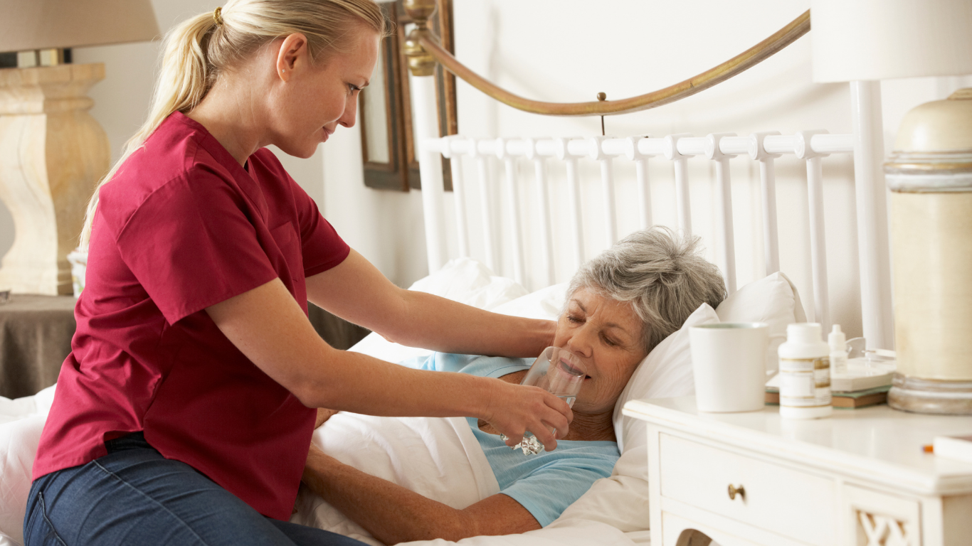 Caregiver in teal scrubs helps seated older adult in bright living room