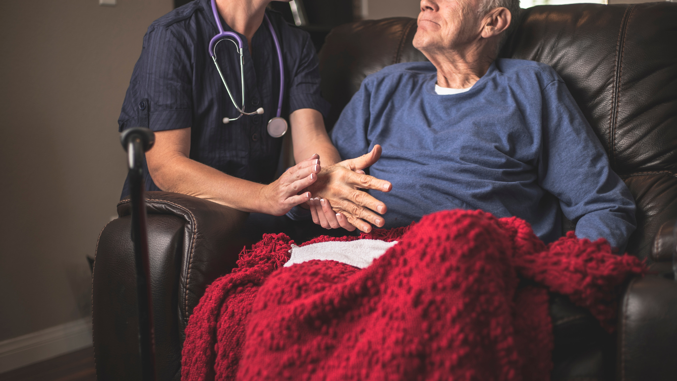 Caregiver in blue scrubs assisting an elderly woman with a walker in a bright home room