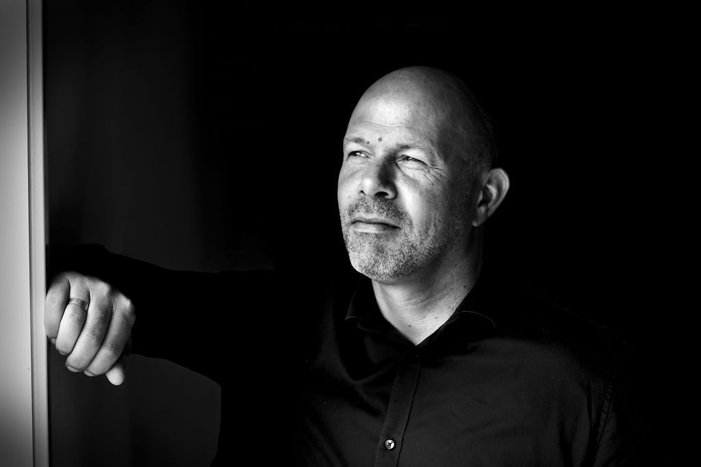 Op een zwart-witportret leunt een persoon tegen een muur en kijkt peinzend weg van de camera.