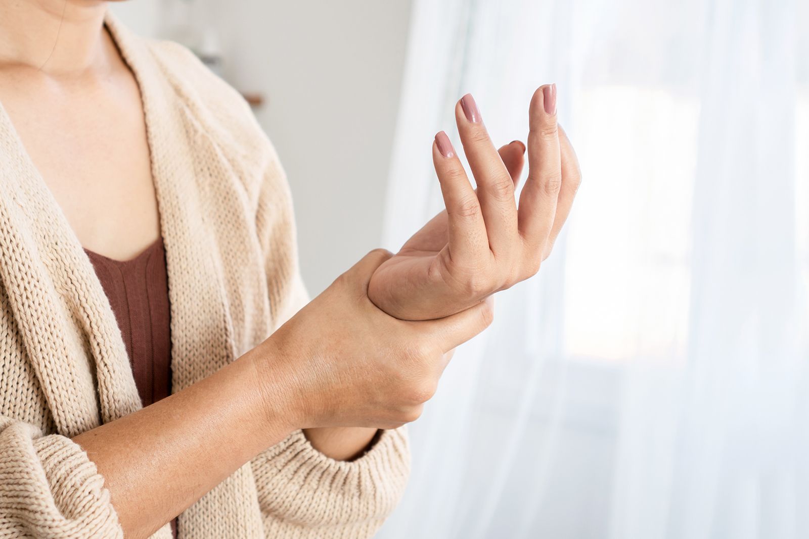 Mujer sujetándose la muñeca, posiblemente debido al dolor, en un espacio interior cerca de una ventana.