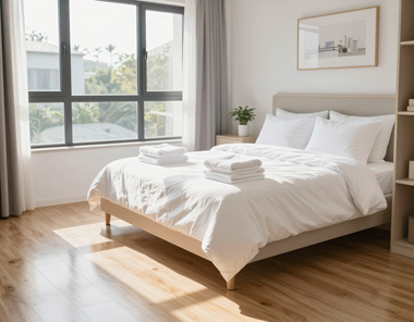 Bright bedroom with a made bed, sunlight through a large window, and wooden floors