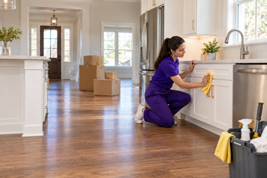 Cleaner in purple uniform wipes kitchen cabinets in a bright home with moving boxes in the background
