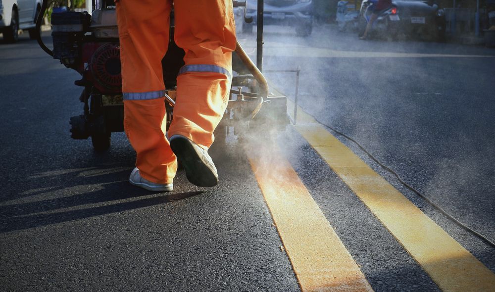 A Man in Orange Pants is Painting Yellow Lines on the Road — Dino's Bobcat–Bitumen & Excavator Hire in Wondai, QLD