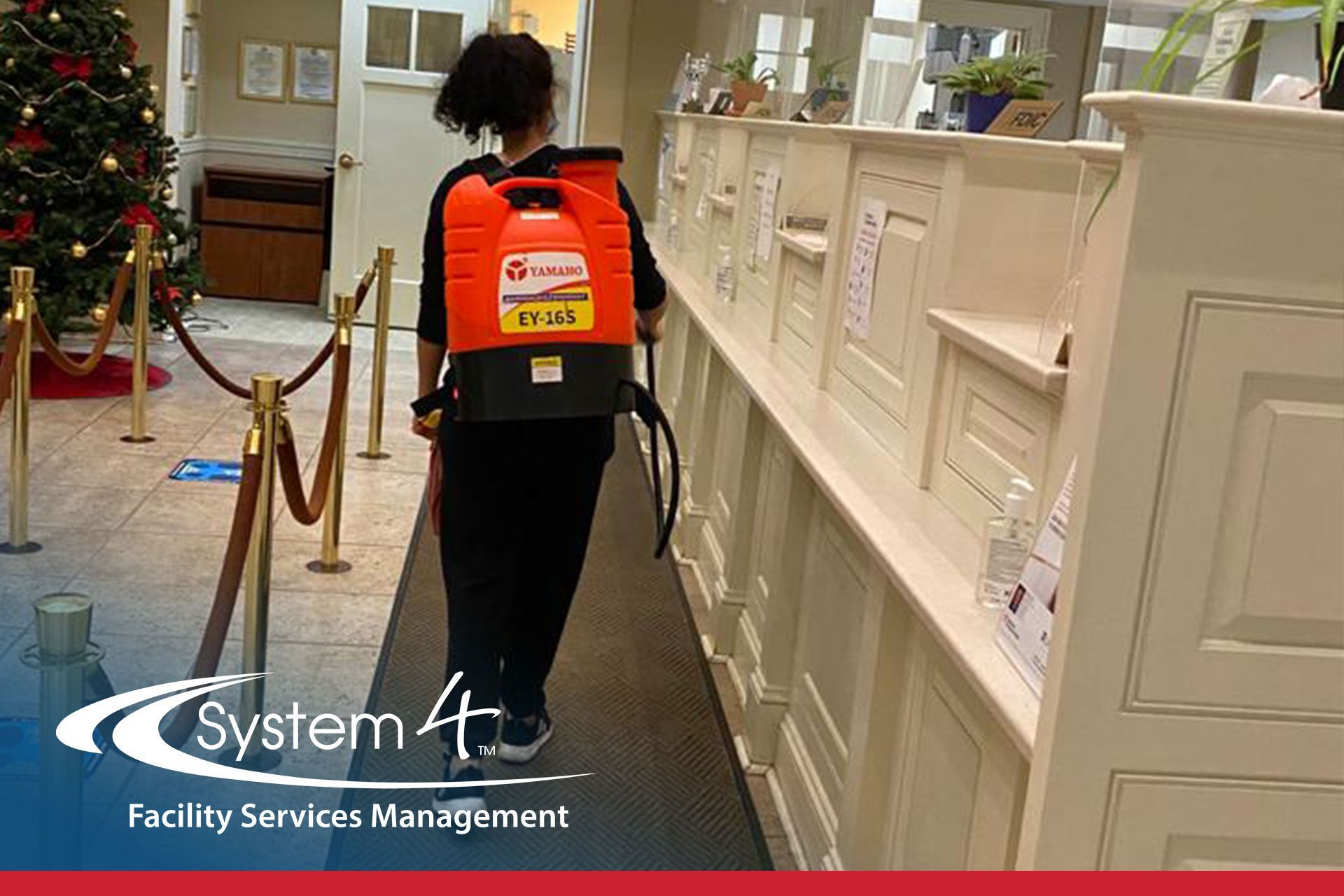 Worker vacuuming a hallway beside a white railing, wearing a red backpack vacuum in a facility lobby