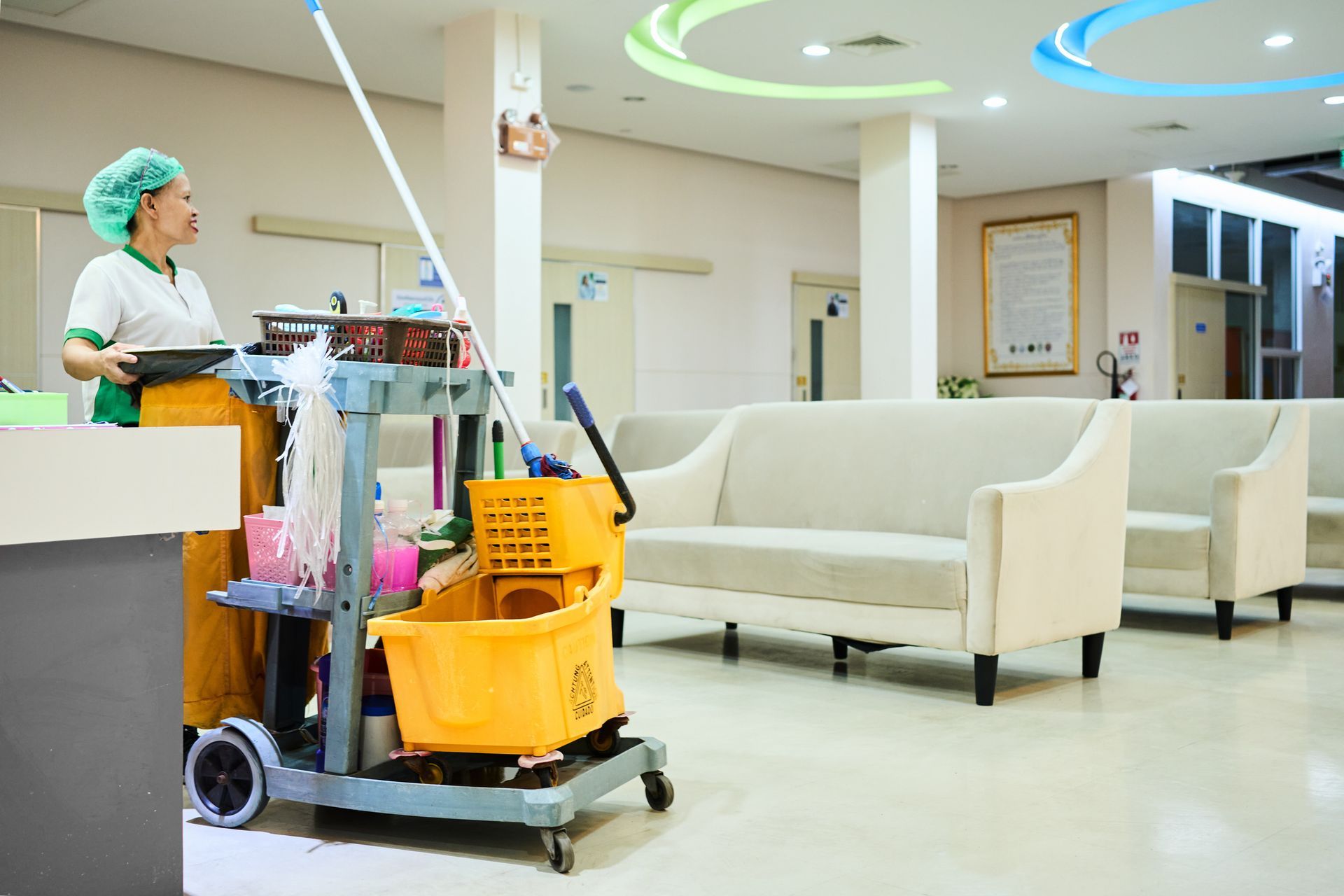 Hospital cleaner mopping a bright waiting room beside white sofas and a yellow cleaning cart