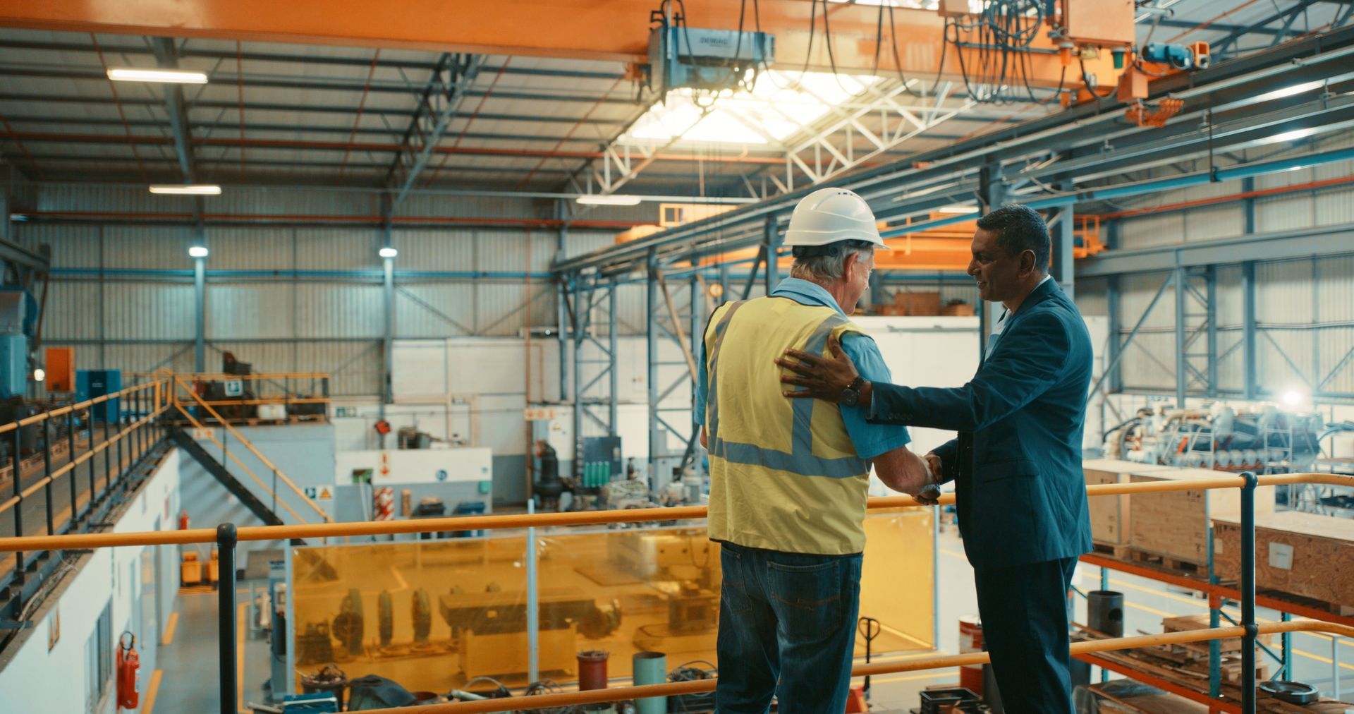 Two workers in hard hats talking on an industrial catwalk inside a factory ceiling area