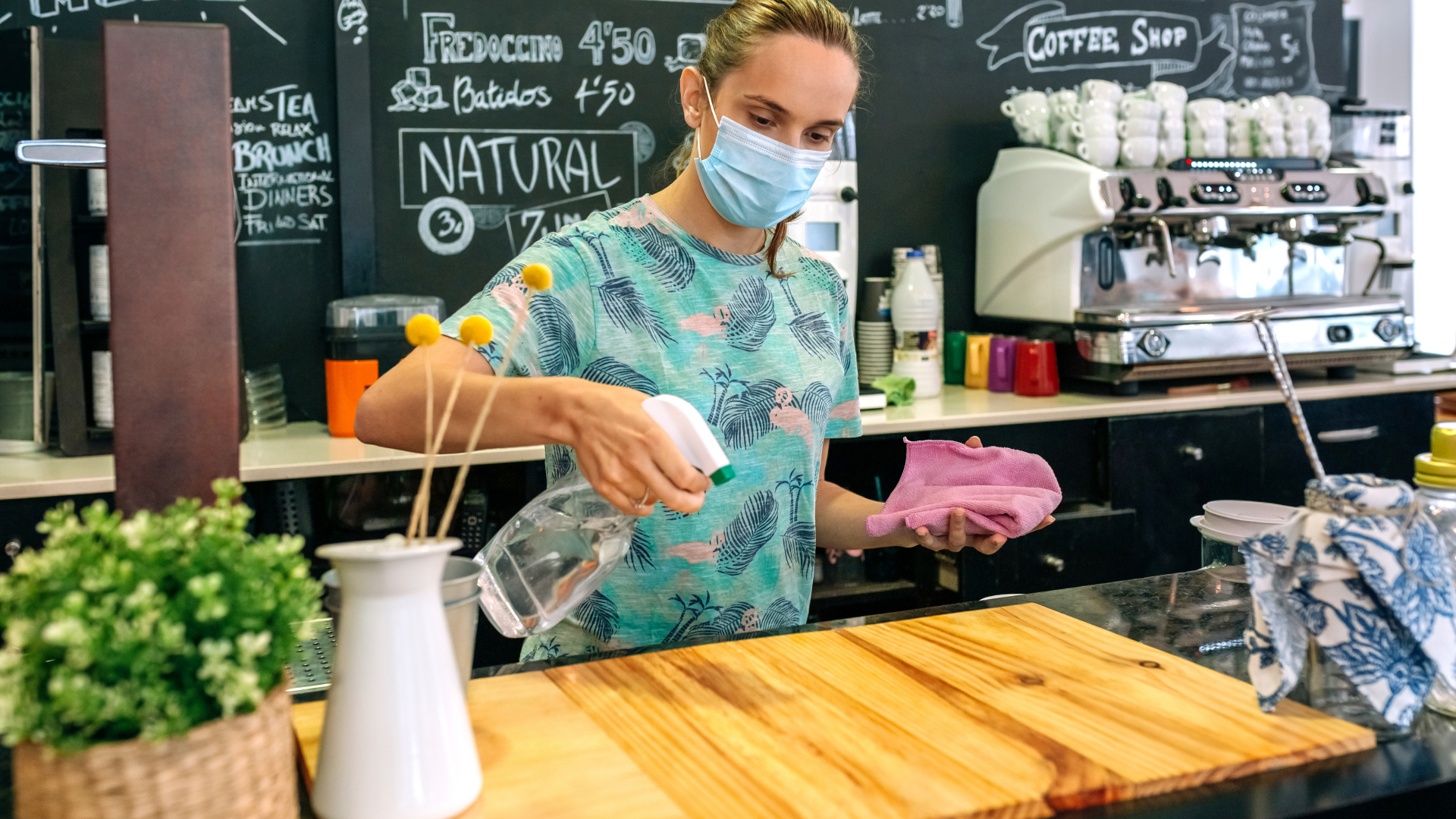 Barista wearing a mask wipes a wooden counter in a café with coffee equipment in the background