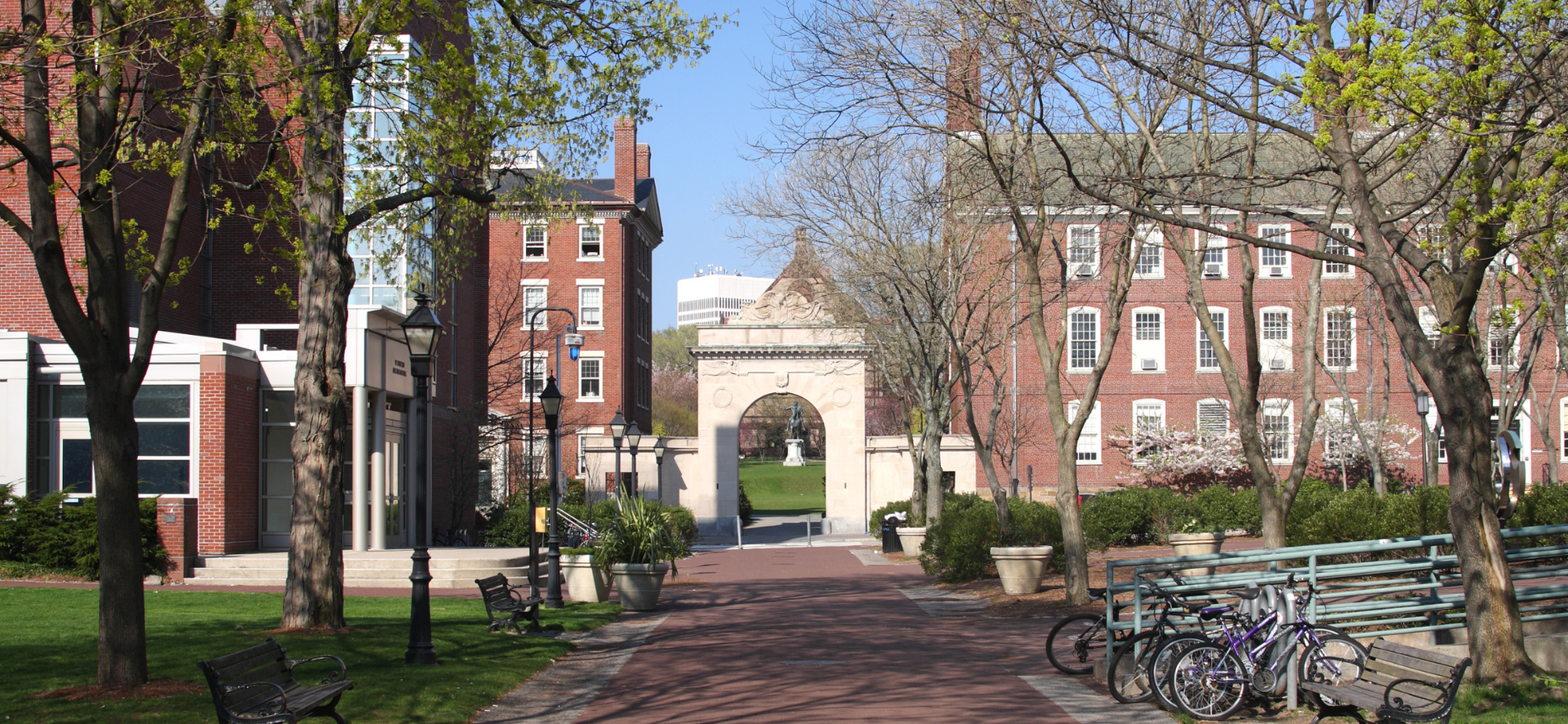 Brick campus walkway between red-brick buildings, with trees, bicycles, and an arched entrance in the distance