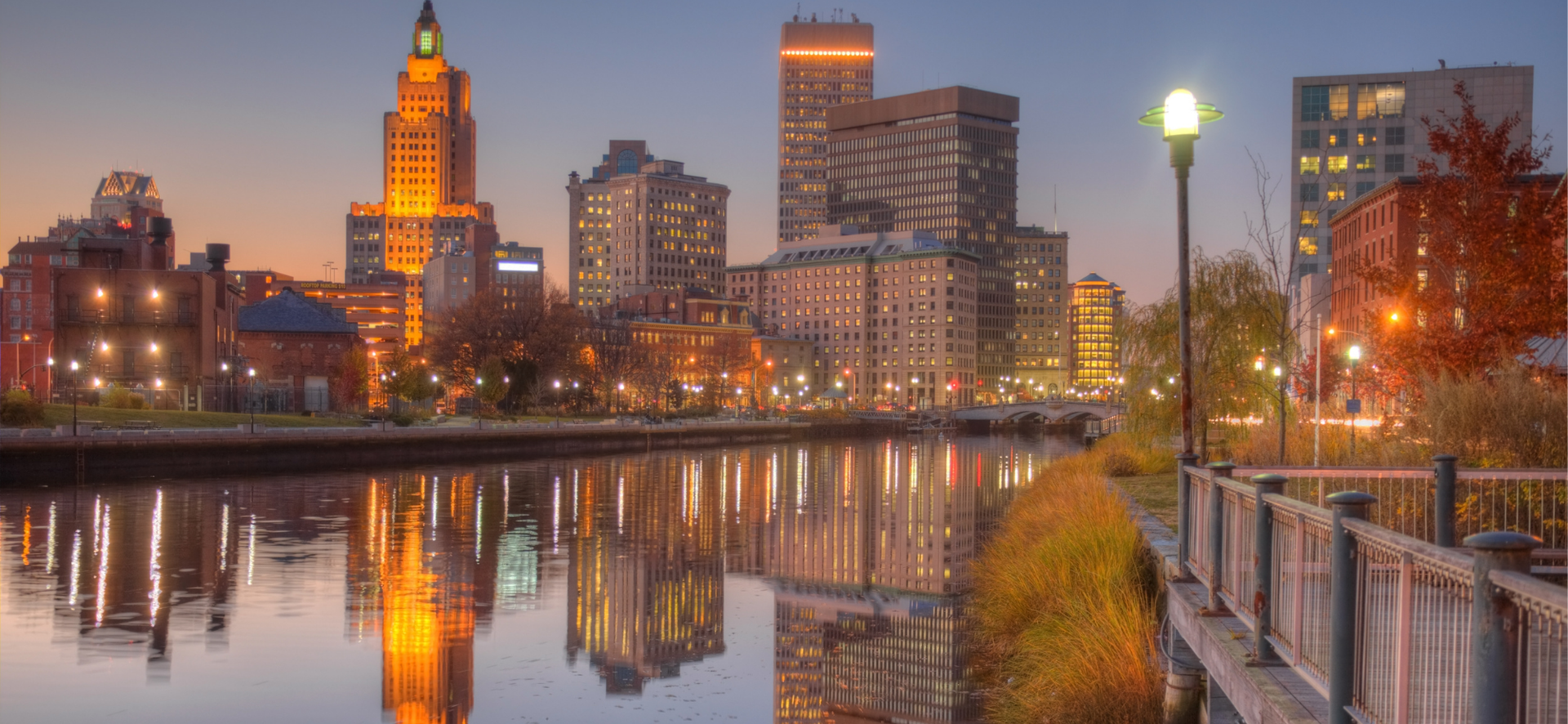 City skyline at sunset reflected in a calm river beside a riverside walkway and autumn trees