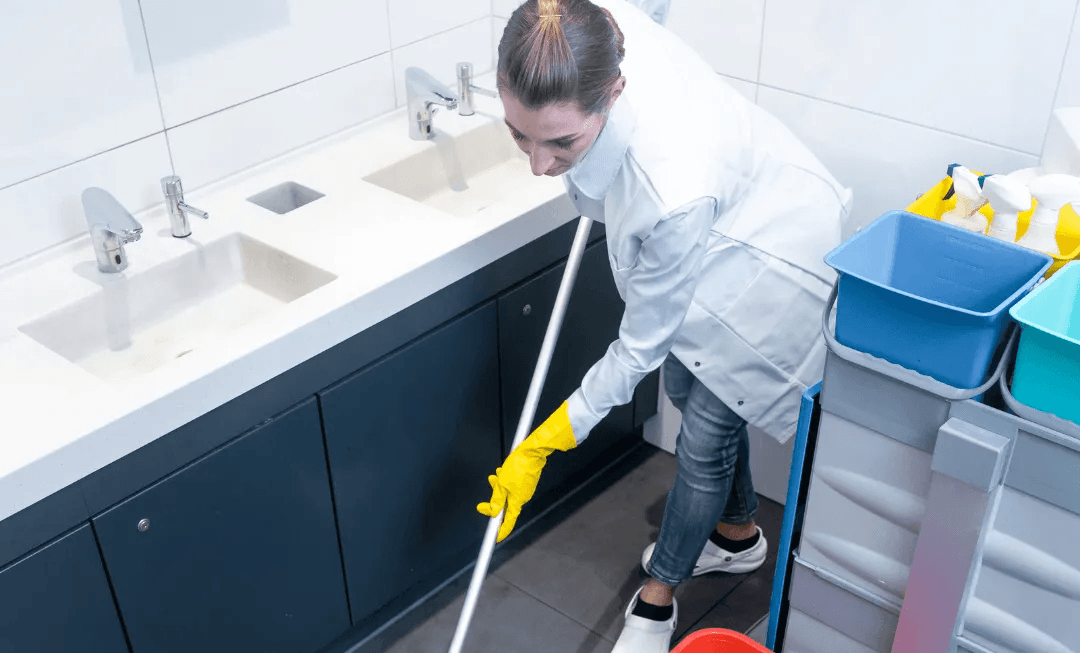 Cleaner mopping a bathroom floor beside sinks, wearing gloves and holding a cleaning cart