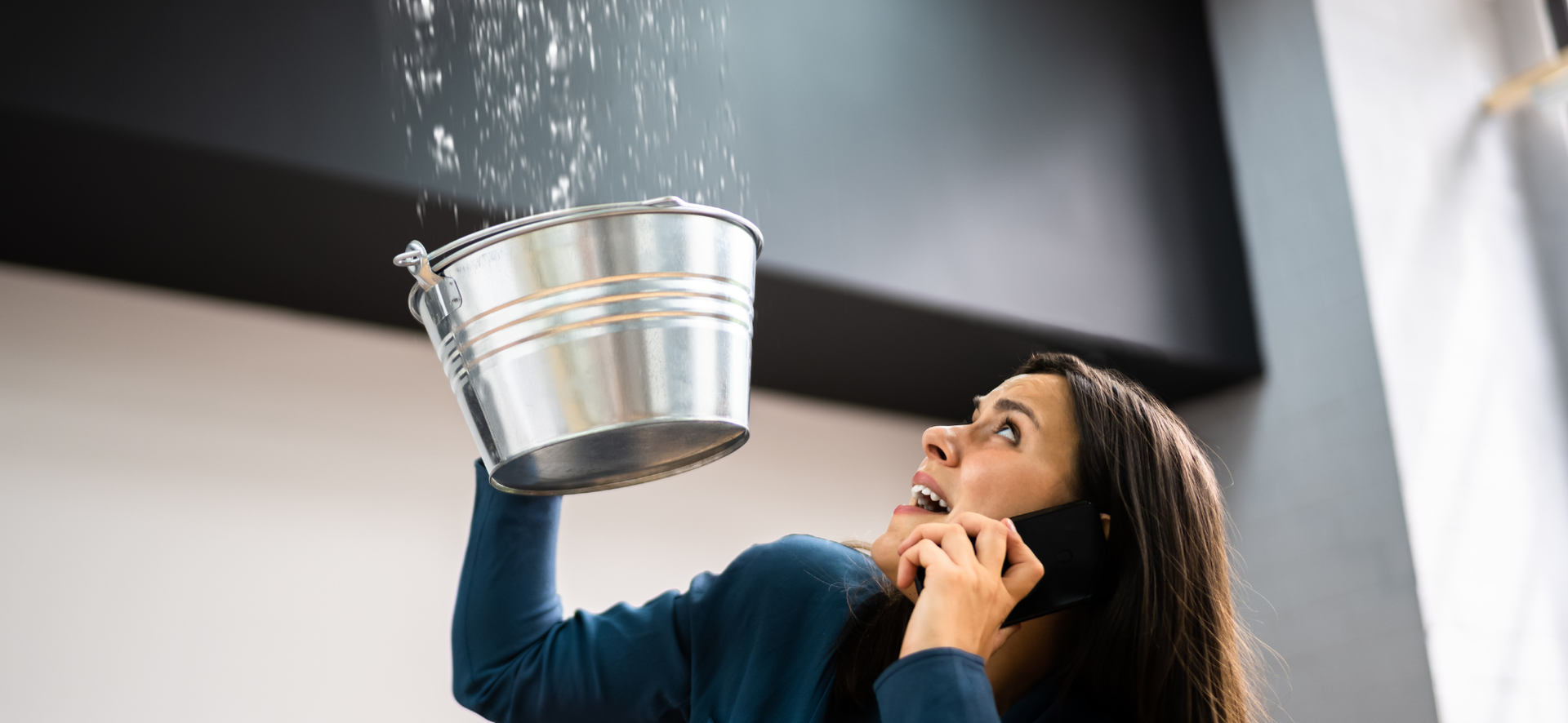 Woman on phone holding a bucket to catch water leaking from the ceiling