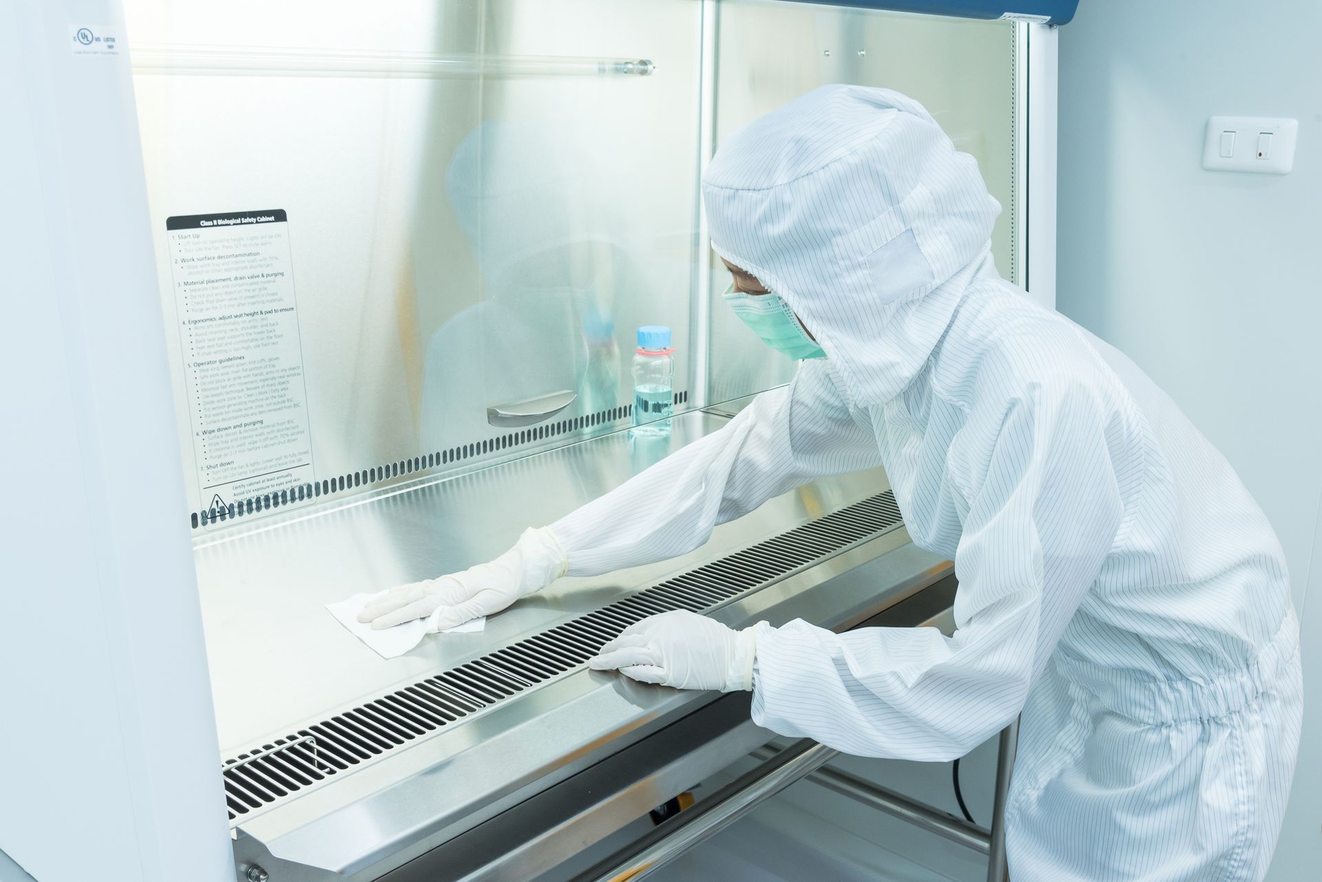 Lab worker in white protective suit cleaning a biosafety cabinet in a sterile laboratory.