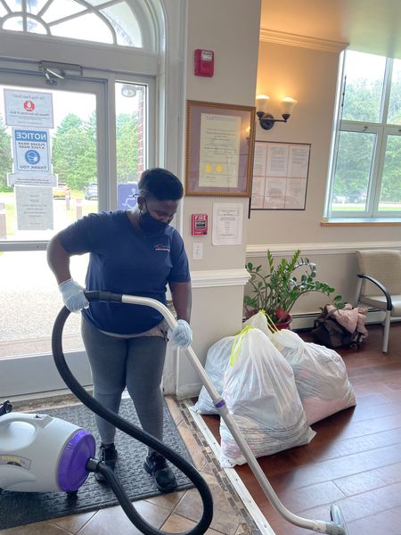 Person vacuuming a lobby floor beside bags and a potted plant near a window.