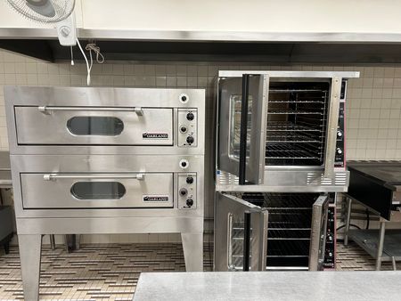 Commercial kitchen with two stainless steel ovens and a proofing rack on a tiled floor.