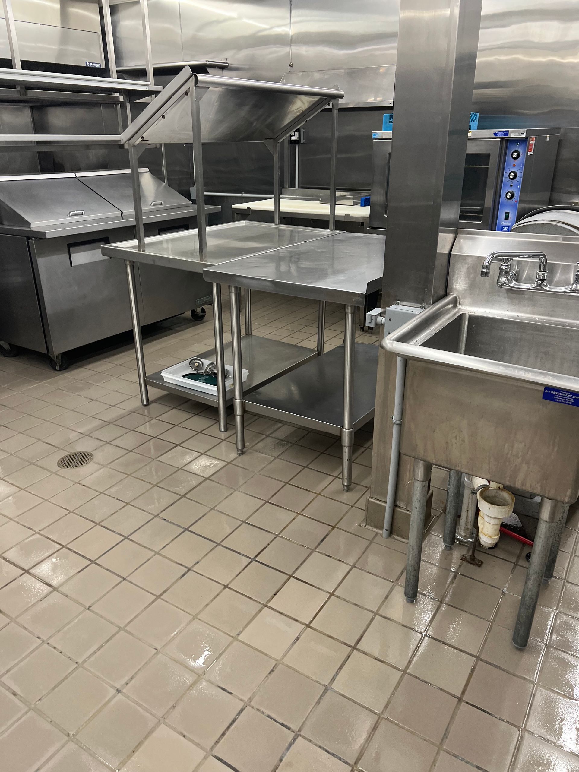 Stainless steel prep tables and a sink in a commercial kitchen with tiled floor.
