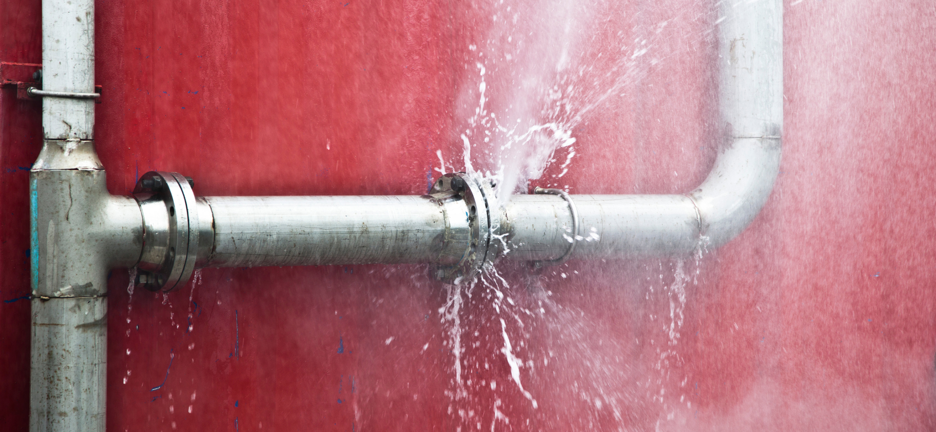 Leaking silver pipe on a red wall, water spraying downward