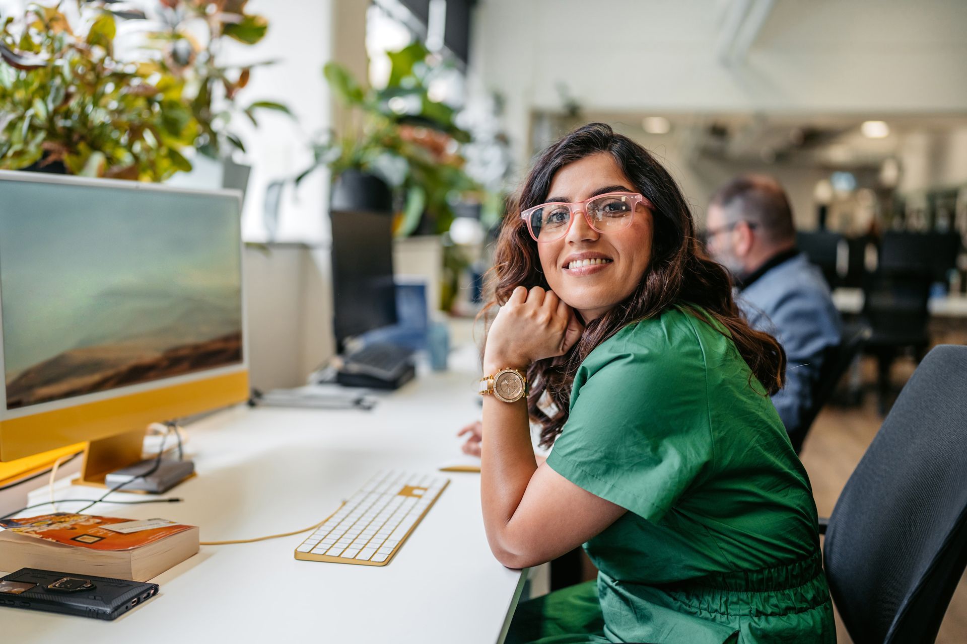 Woman smiling at a desk in a bright office, with a computer monitor and coworkers in the background