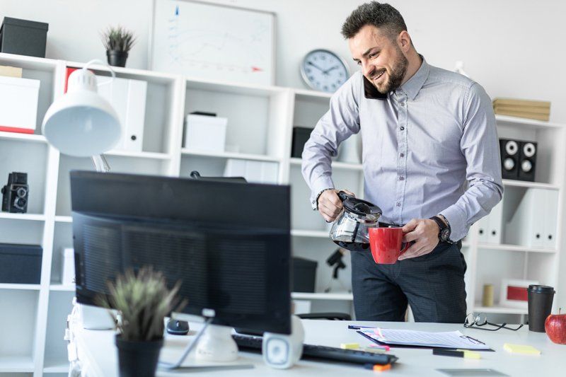 Man standing at an office desk, holding a red mug and looking at a computer monitor.