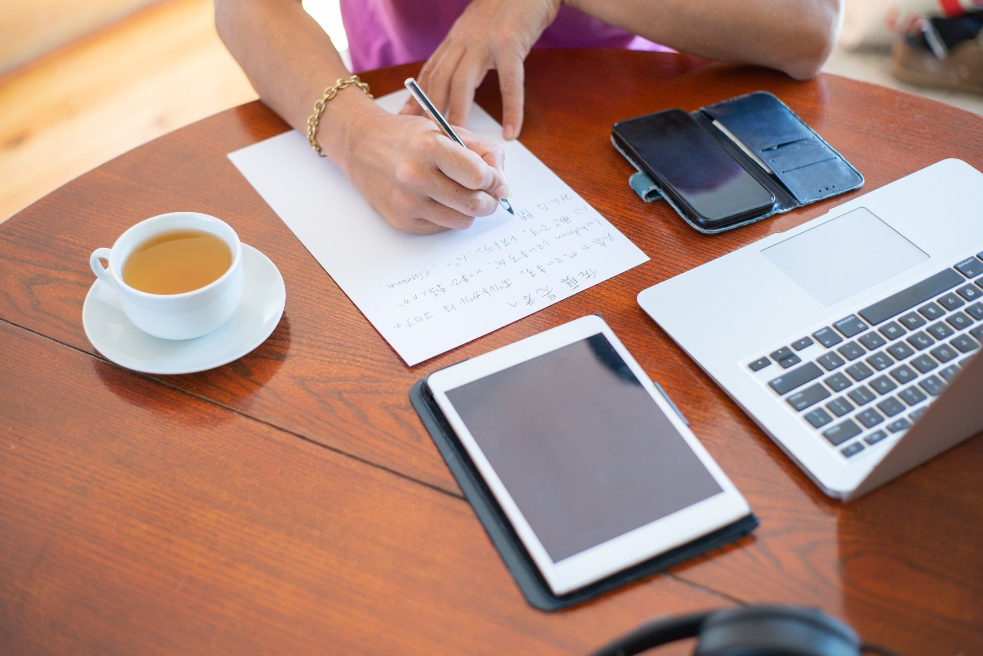 Hands working over papers at a desk with a laptop, tablet, phone, and cup of coffee
