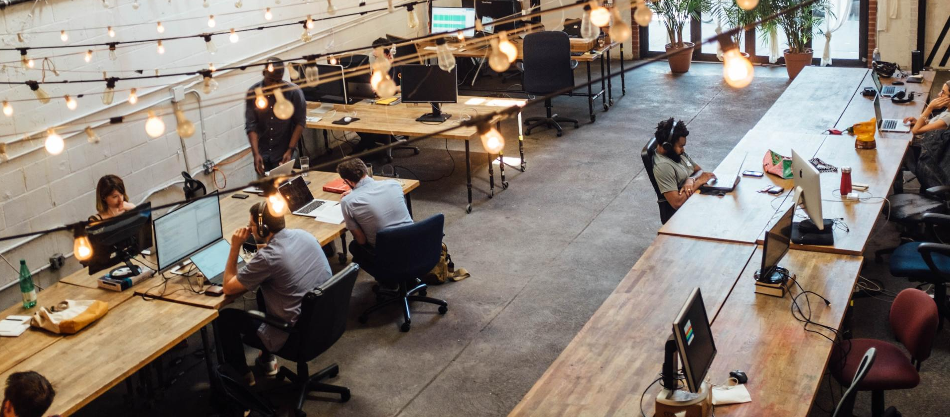 Open-plan office with people working at long wooden desks under hanging lights