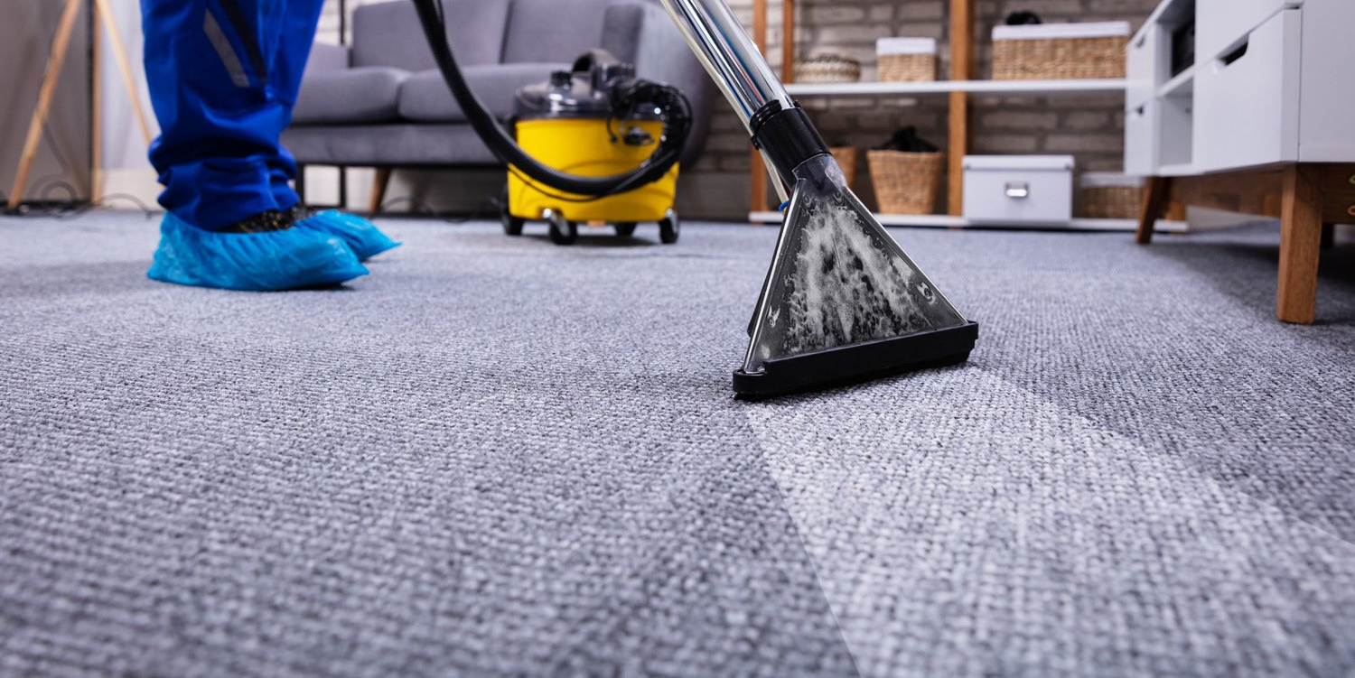Mop cleaning a gray carpet beside a yellow vacuum in a room with blue-capped feet visible