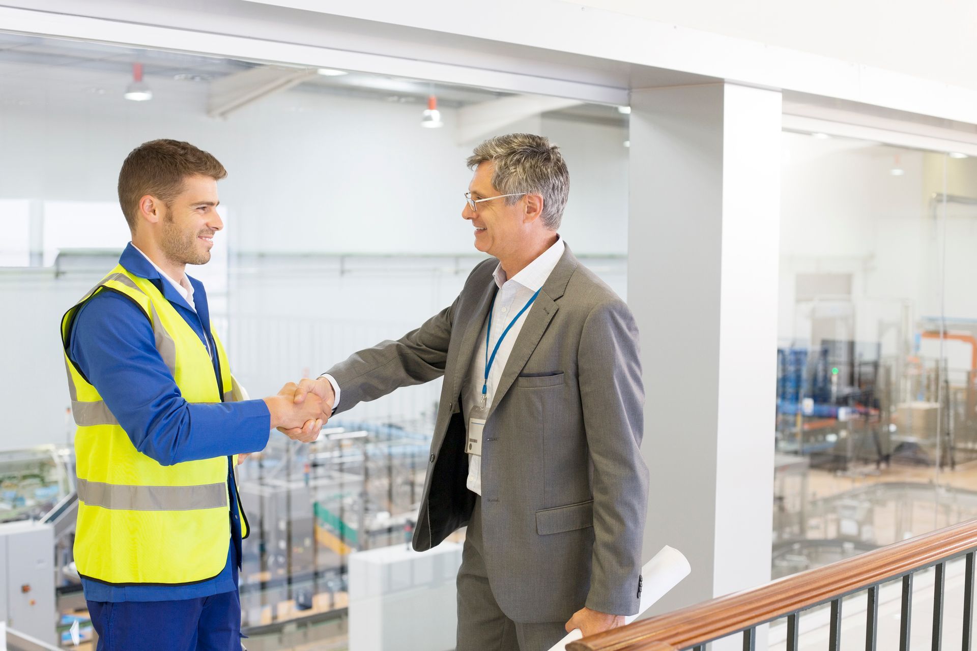 Two men shake hands in a bright office, one in a yellow safety vest and one in a gray suit.