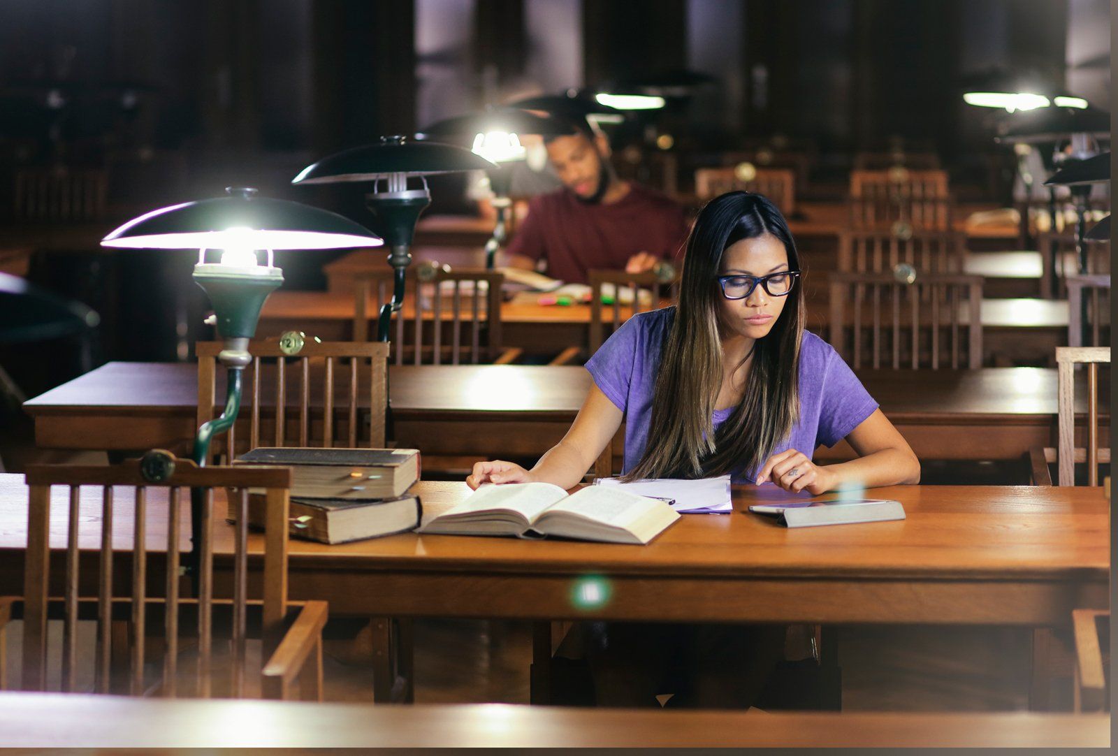 Student studying at a library table under warm lamp light, reading an open book.