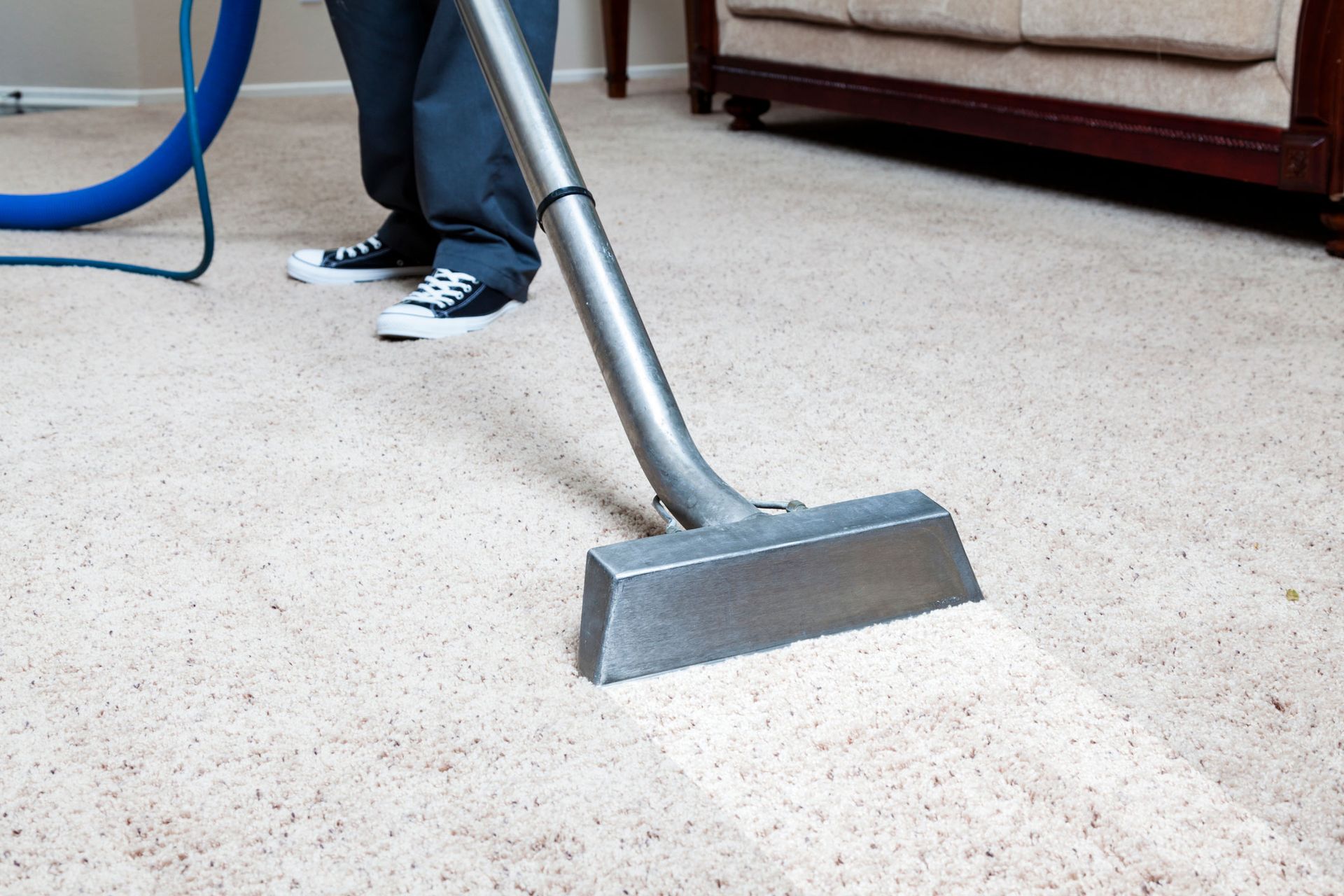 Vacuum cleaner head on beige carpet near a couch and a person’s shoes
