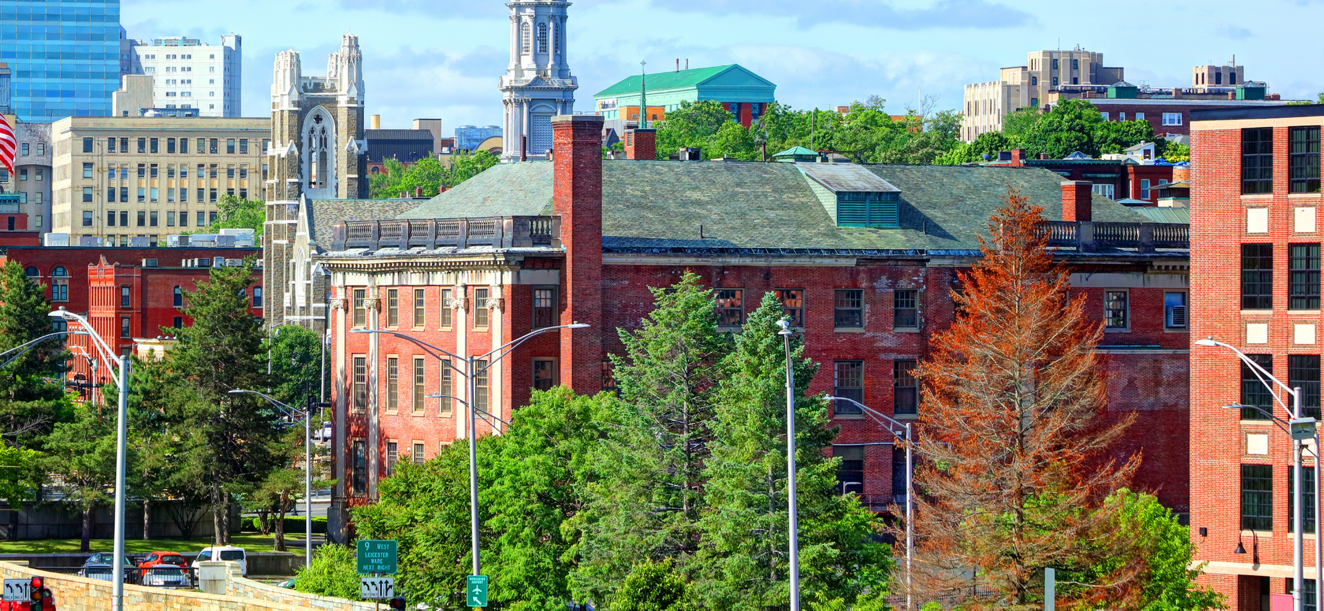 City skyline with historic brick buildings, trees, and a green-roofed structure under a blue sky