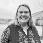 Smiling woman with glasses in a patterned blouse standing outdoors near a bridge