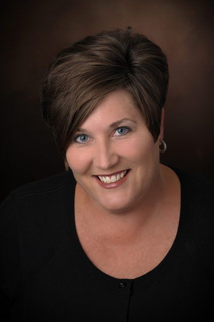 Woman with short brown hair, smiling, wearing black shirt, against a dark brown background.