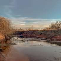 A river flows through a landscape, brown water, trees, red-brown soil, and a cloudy sky.