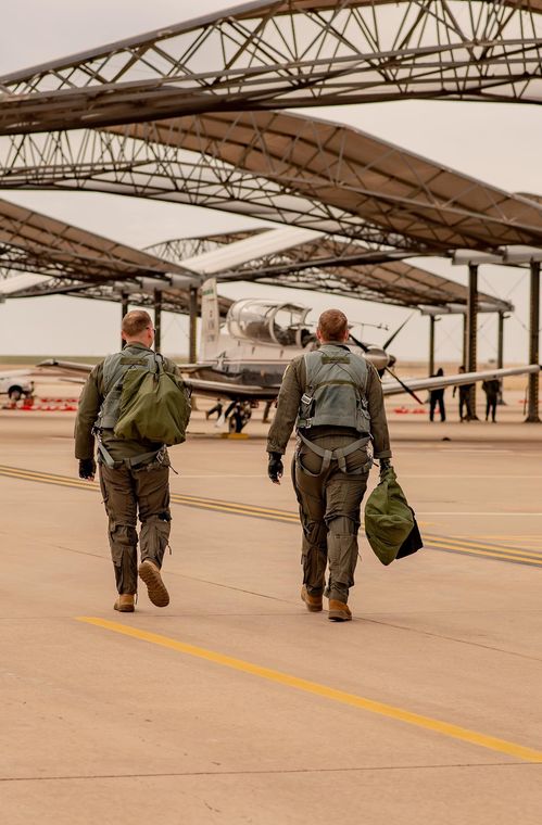 Two people in flight suits walk towards a propeller plane on a tarmac under a hangar.