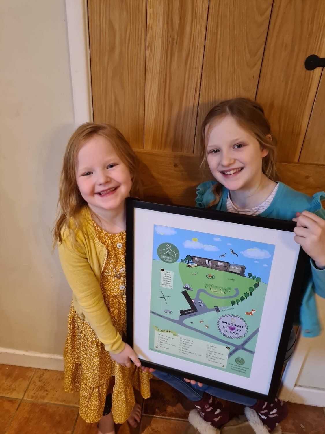 Two smiling girls holding a framed map illustration. One wears yellow, the other blue. Wooden door background.