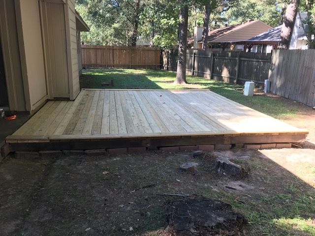 Raised wooden deck in a backyard beside a house, with a fenced yard and trees in the background