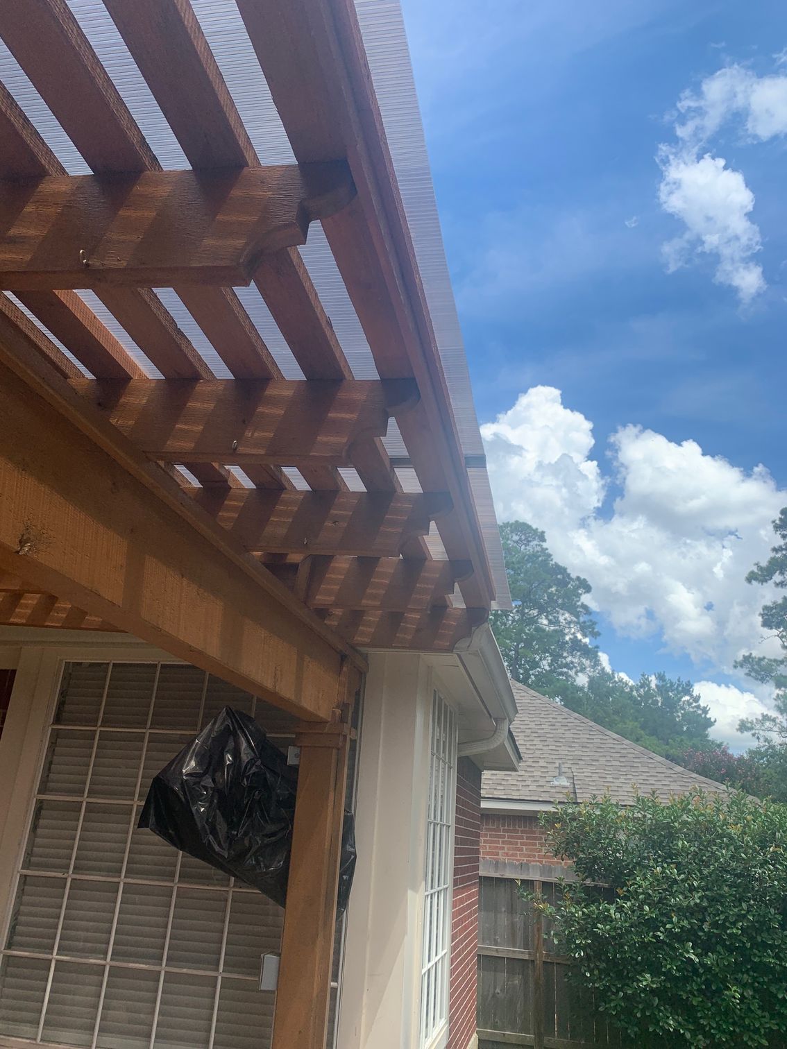 Backyard patio with wooden pergola, brick wall, hanging black bag, and blue sky with clouds