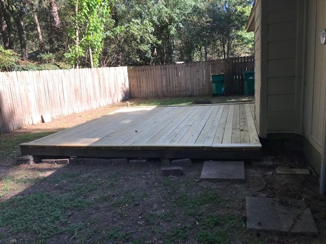 Wooden backyard deck under construction beside a house, with a fenced yard and trees beyond.