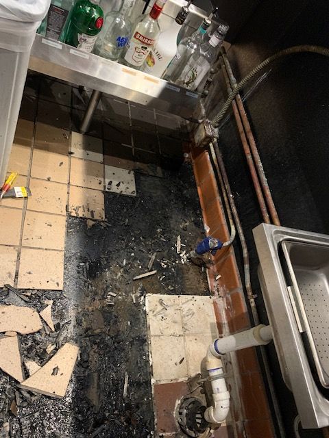 Burnt, charred kitchen floor beside a sink and counter with scattered debris and damaged tiles