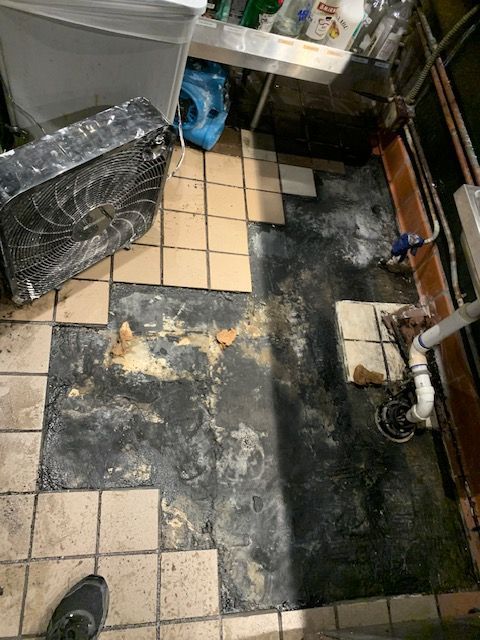 Water-damaged floor with torn black vinyl, exposed beige tiles, and a wet, messy basement corner