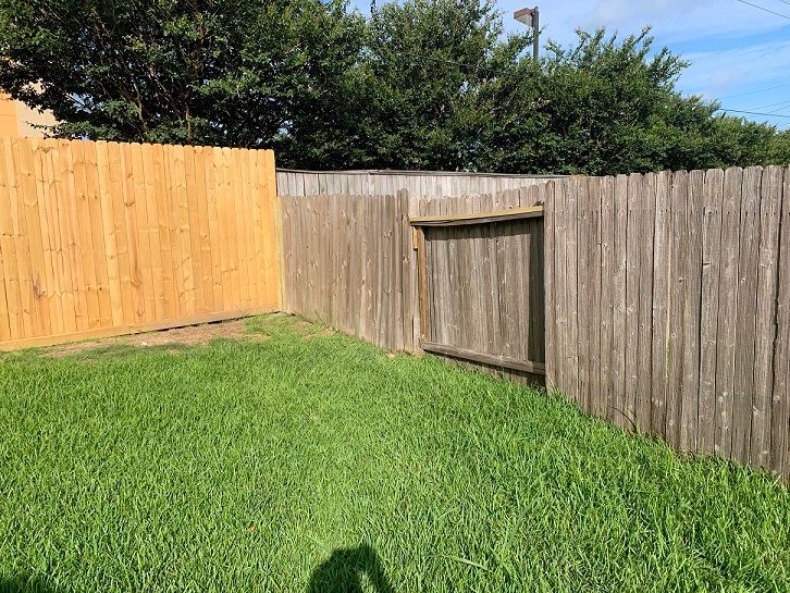 Backyard corner with green grass, a wooden fence, a gate, and trees beyond the fence